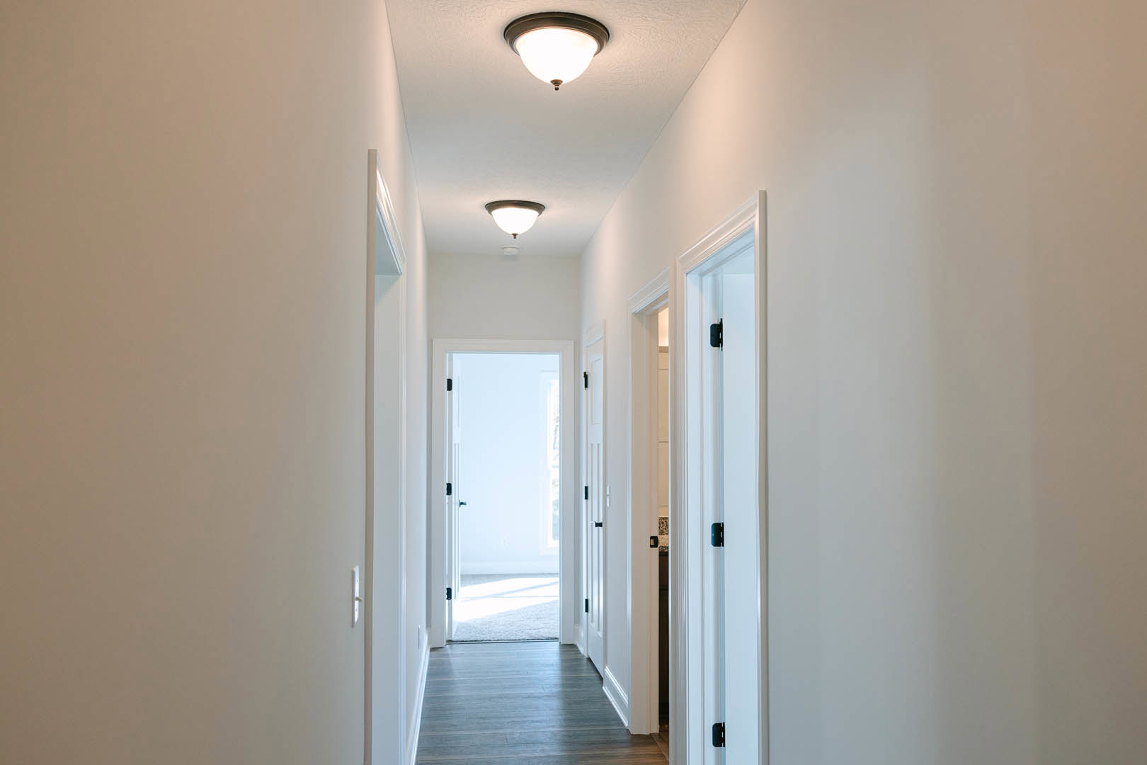 Hallway with smooth white walls, recessed ceiling lights, hardwood flooring, and a white door illuminated by natural light