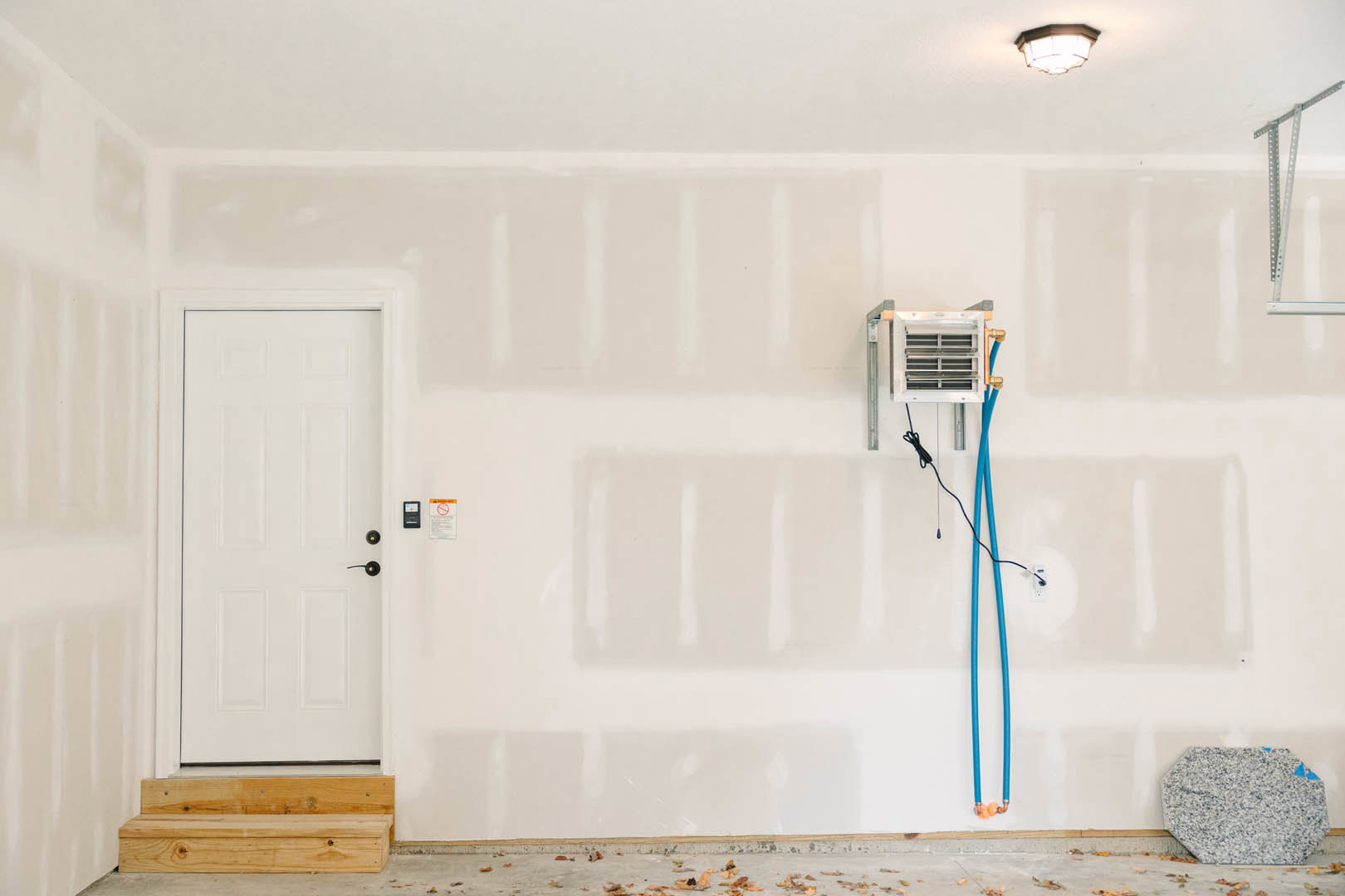 White plaster wall with two white doors, one featuring black knobs and blue hoses; wood step and blue-and-white speckled tile flooring visible.