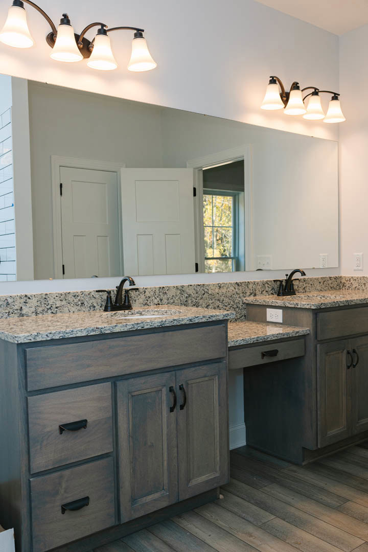 Granite bathroom countertop with undermount sink, large framed mirror, white door with black handle, window overlooking trees, dual bulb light fixture above mirror, tile flooring