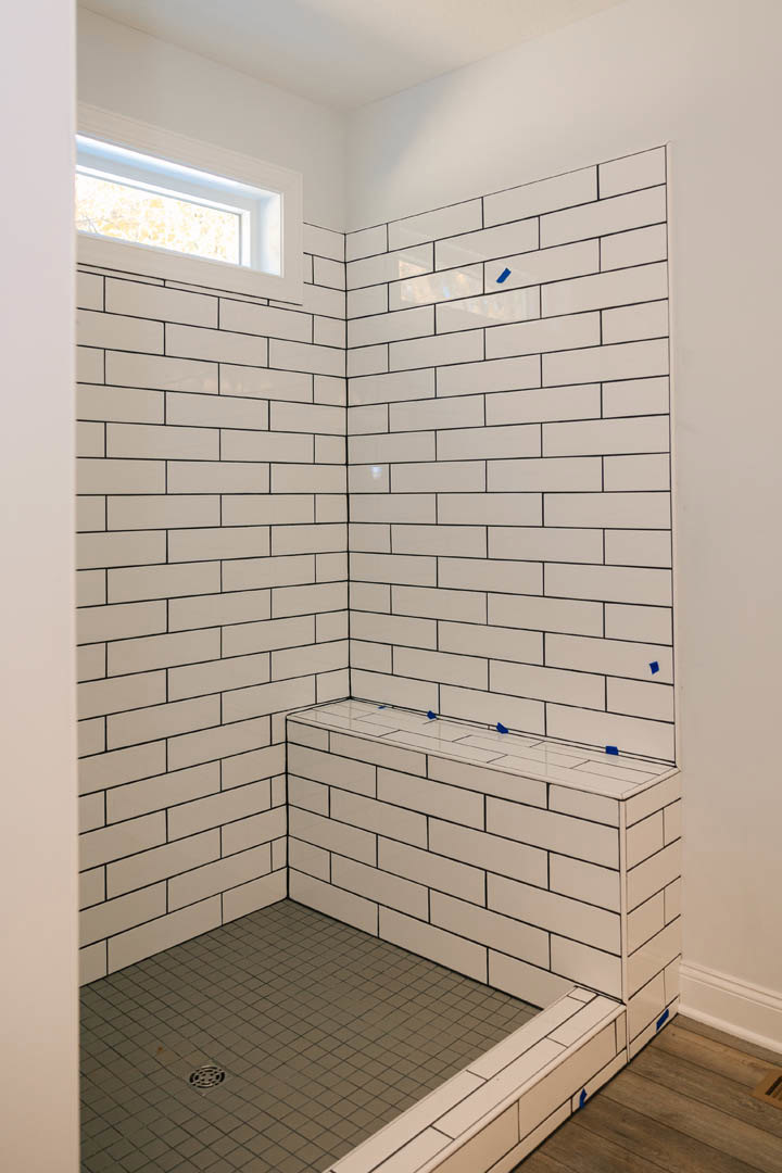 White tile shower with built-in bench, white tile floor, wood flooring with blue tape, window, and white tile wall marked with blue tape