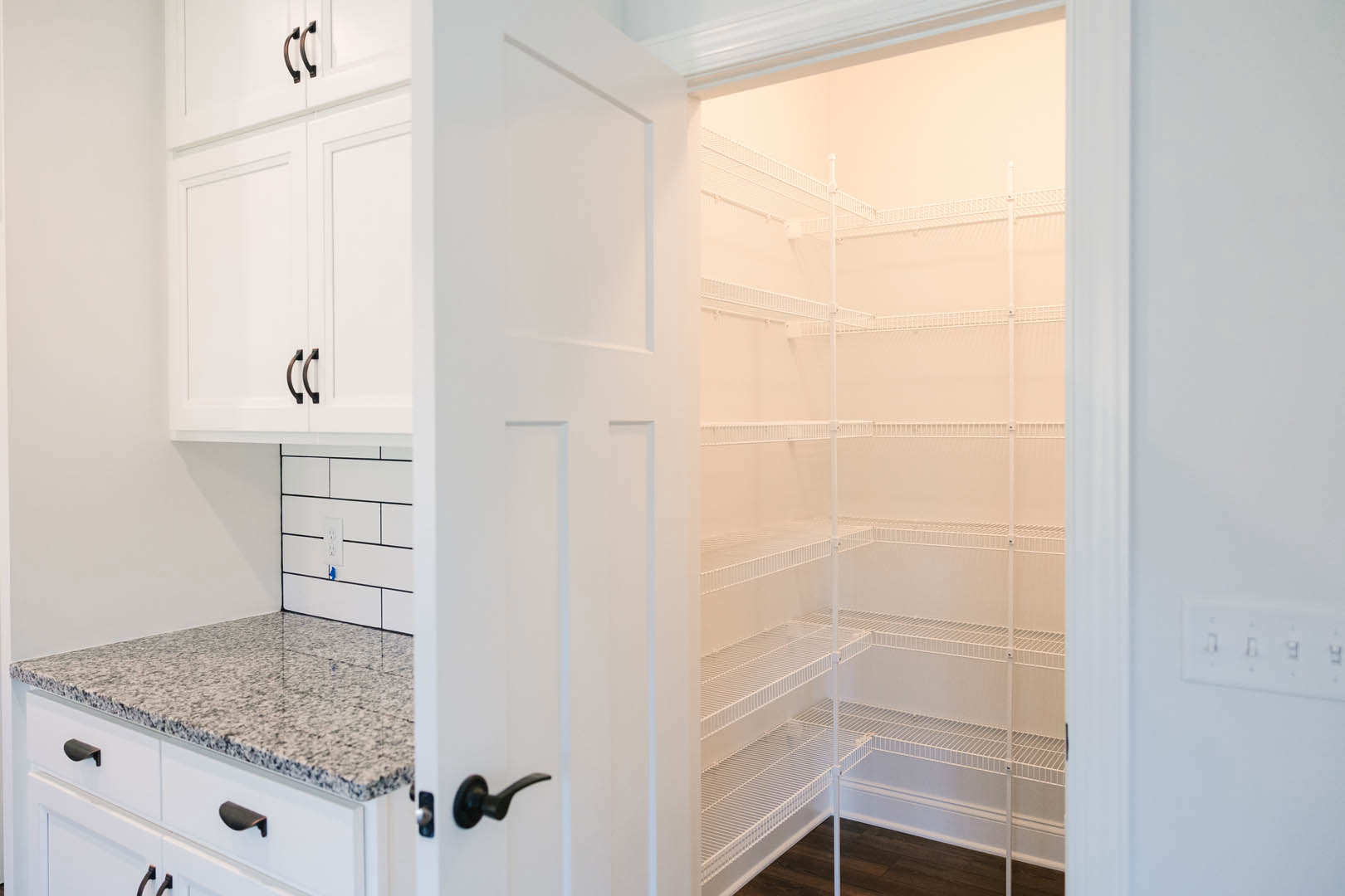Walk-in pantry with white shelves, light stone countertop, white cabinetry, and stainless steel door handle