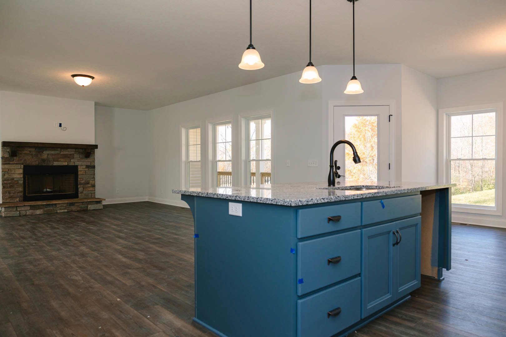Blue kitchen island with drawers and sink, wood flooring, white outlet on blue wall, black-framed fireplace with glass window, close-up of chrome faucet, white cabinetry and