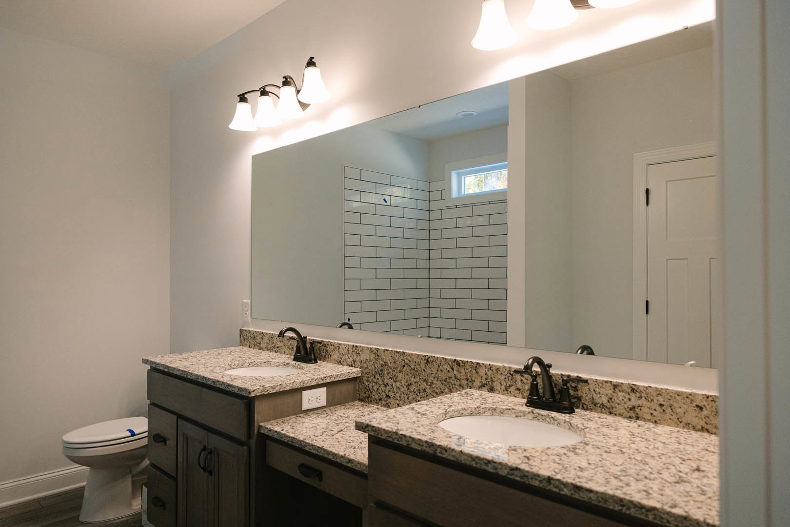 Bathroom with a wide framed mirror above a white countertop, chrome faucet and sink, four-light fixture, toilet marked with blue tape, light gray tile walls, and dark wood