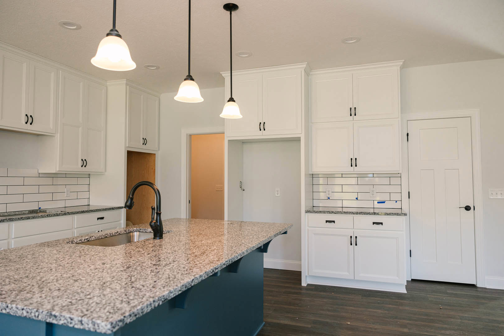White kitchen cabinets with black handles, granite island countertop featuring an undermount sink and modern faucet, white door with black handle, neutral wall finish, stainless