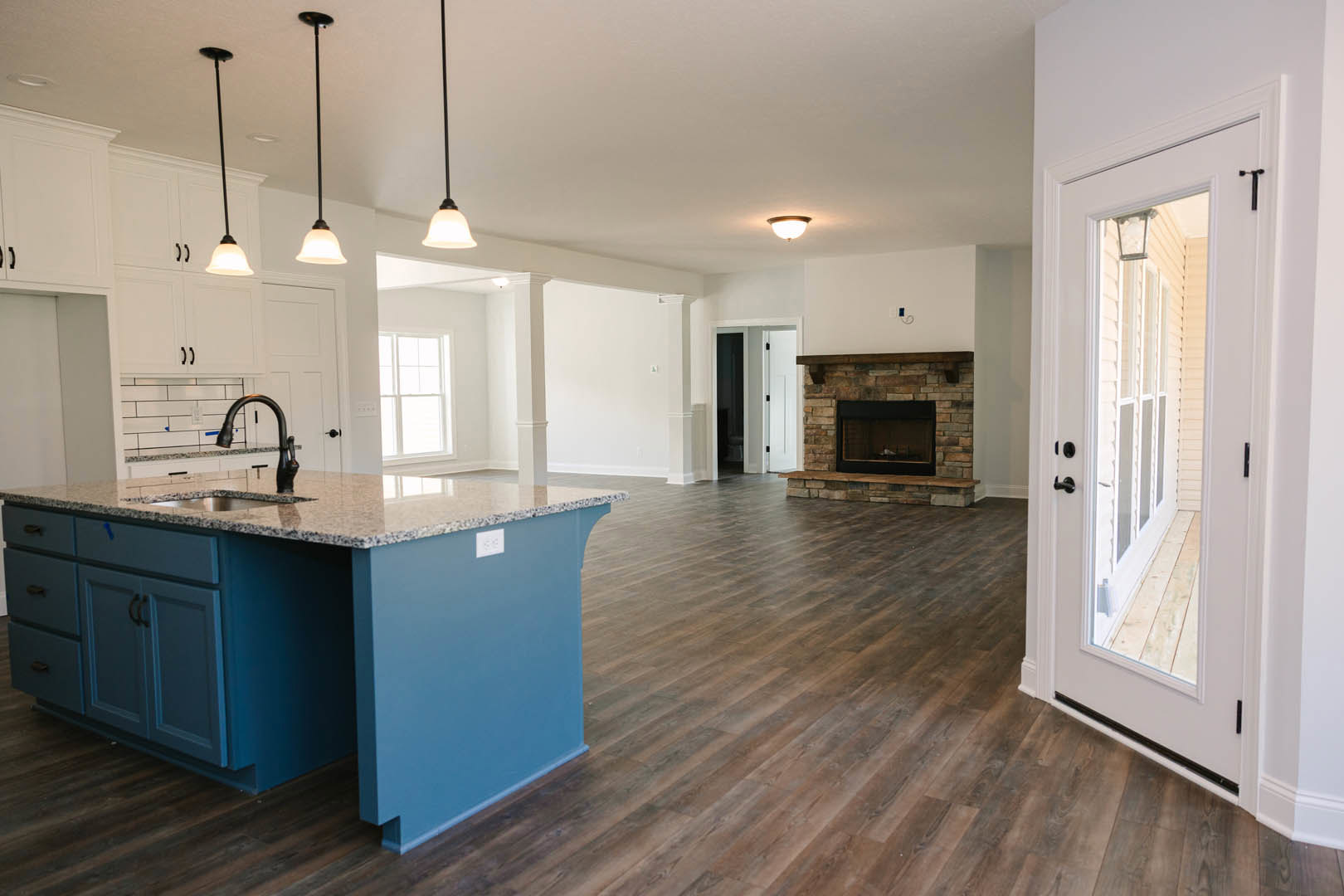 Open kitchen and living room featuring wood flooring, central kitchen island with built-in sink, white cabinetry, fireplace with glass door, white door with glass panels, and black