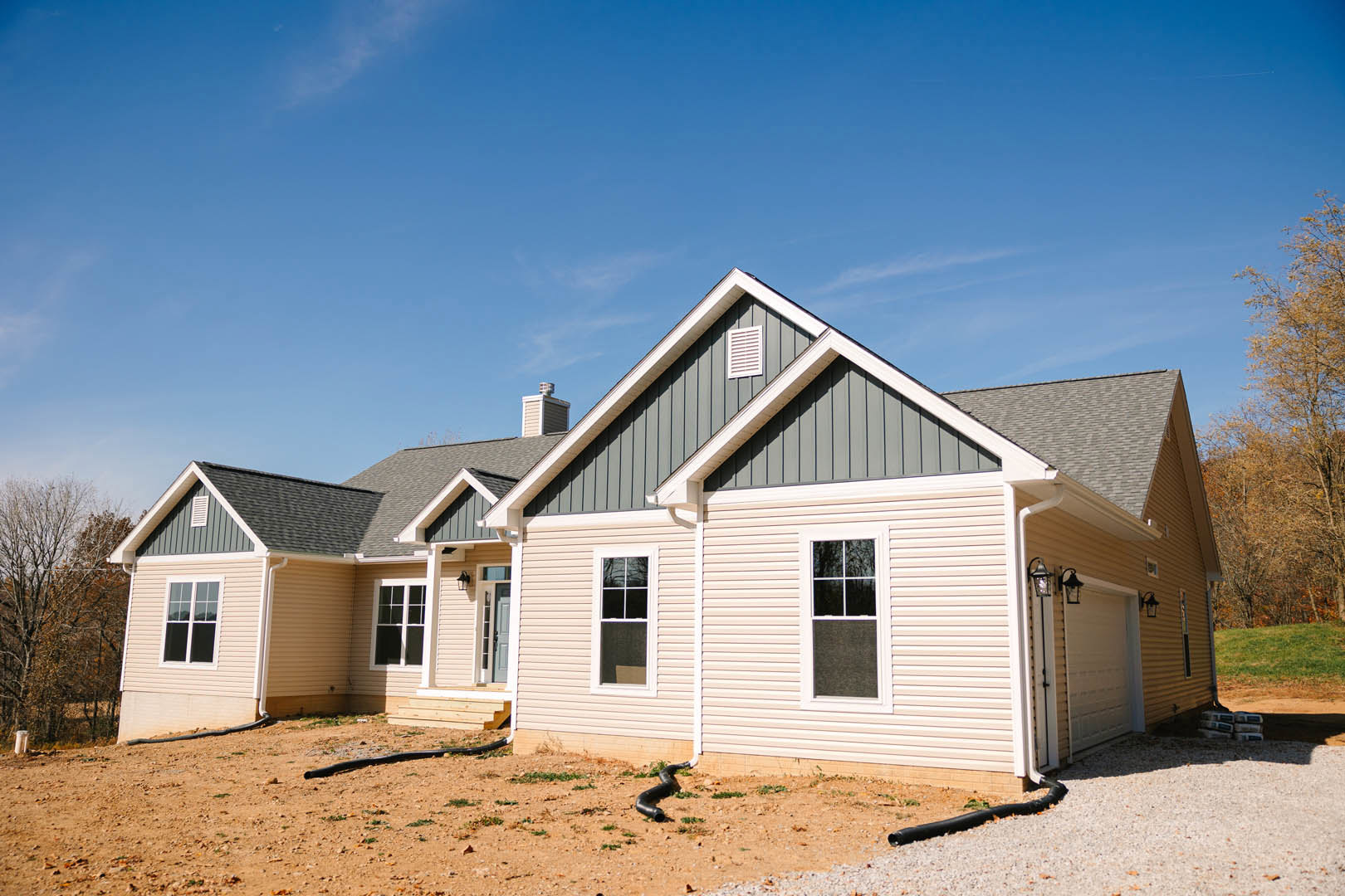 Two-story house with gray siding, white-framed windows, and brick chimney, surrounded by leafless trees, gravel driveway, and black pipe lying near the foundation.