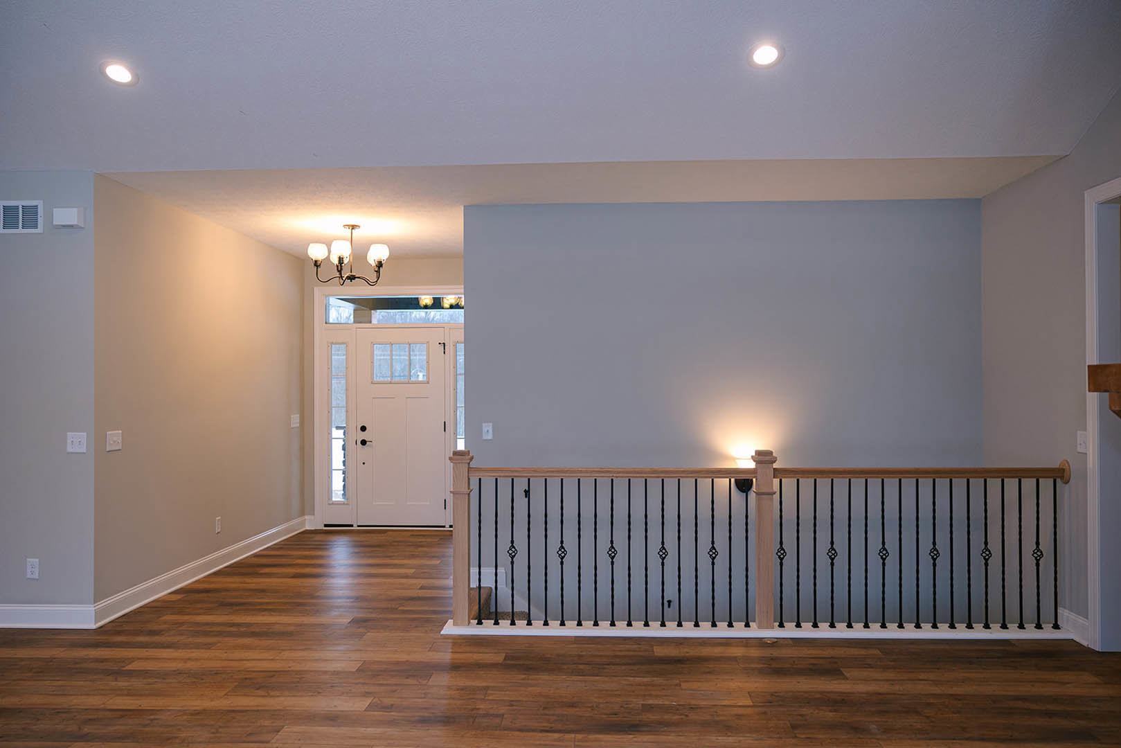 Hallway with light wood flooring, black metal railing, white door with glass panes, white walls, ceiling chandelier, and recessed lighting