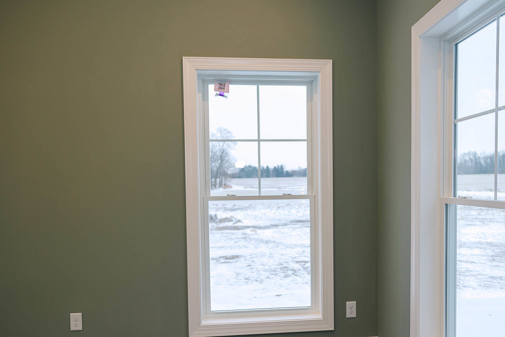 Large window with white trim overlooking snowy field, paper tag taped to glass, green wall with white electrical outlet visible indoors