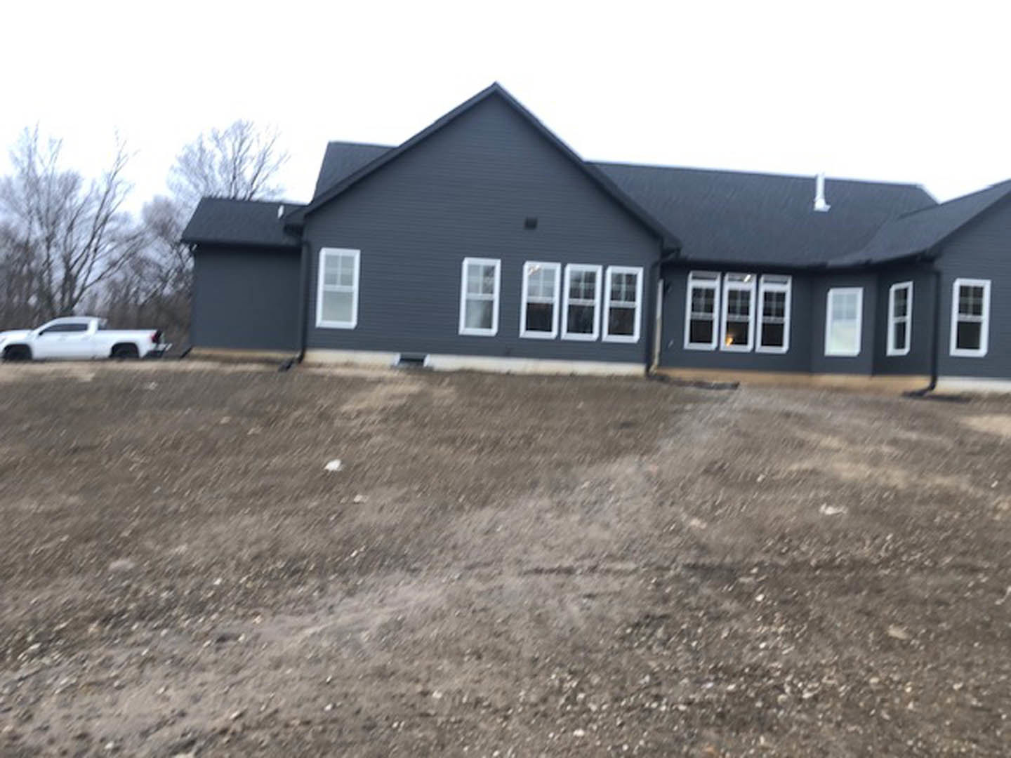 Two-story house with multiple windows, light-colored exterior, surrounded by a dirt field, sparse landscaping, and a white car parked nearby