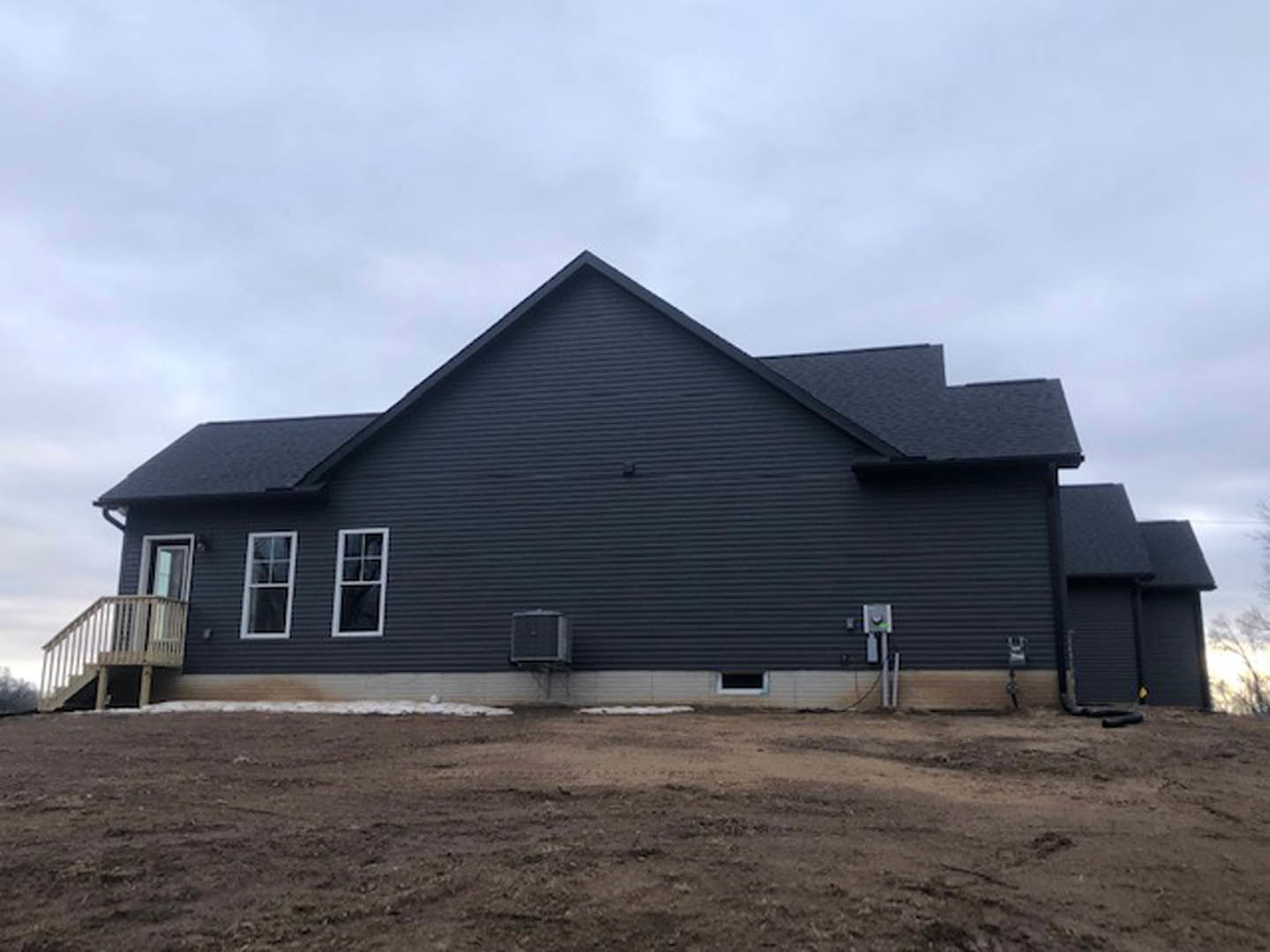 Black house with white siding and window frames, white sign near entrance, dirt yard in foreground, cloudy sky overhead