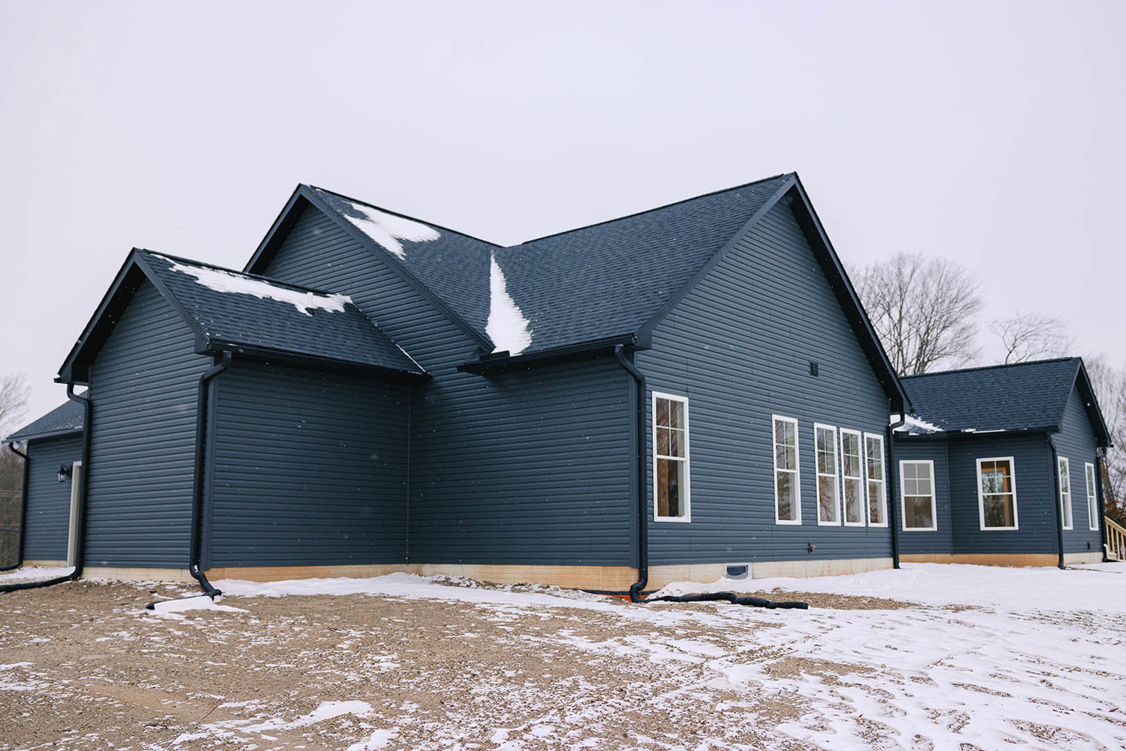 Two-story house with white-framed windows, snow covering the roof and ground, leafless tree in front yard, exposed pipe near foundation