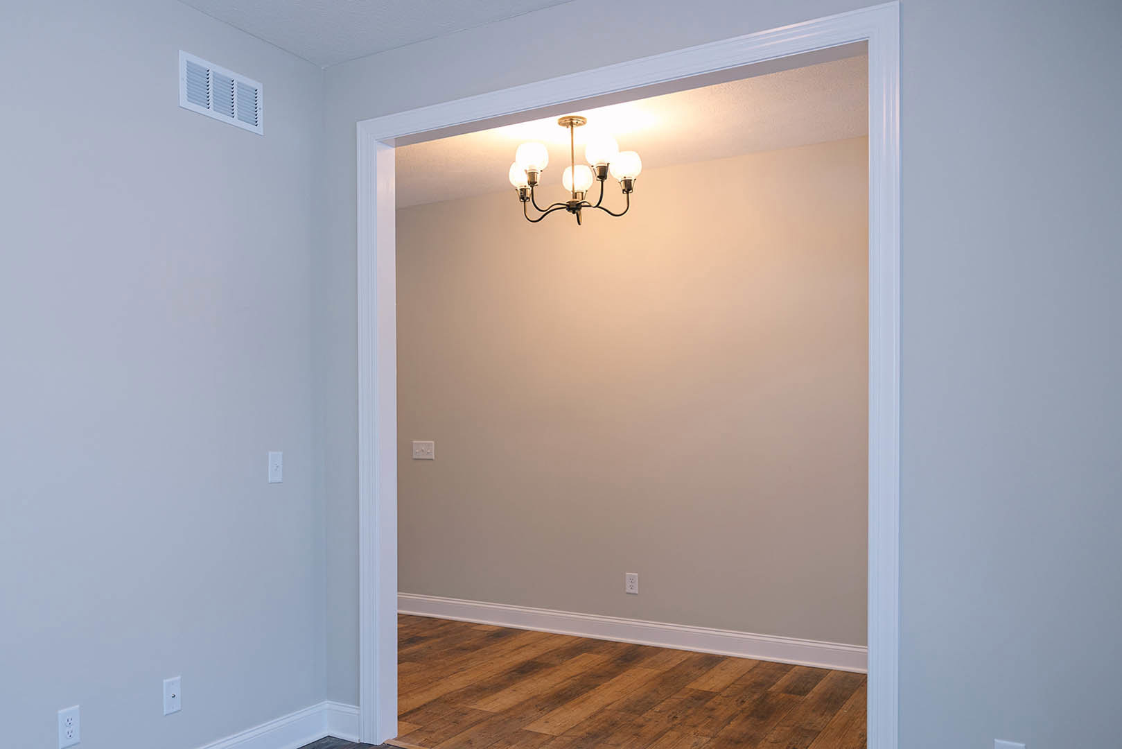 Brushed metal chandelier with exposed bulbs hanging in hallway, wooden floor with white baseboard, white wall vent, plaster walls, white door molding