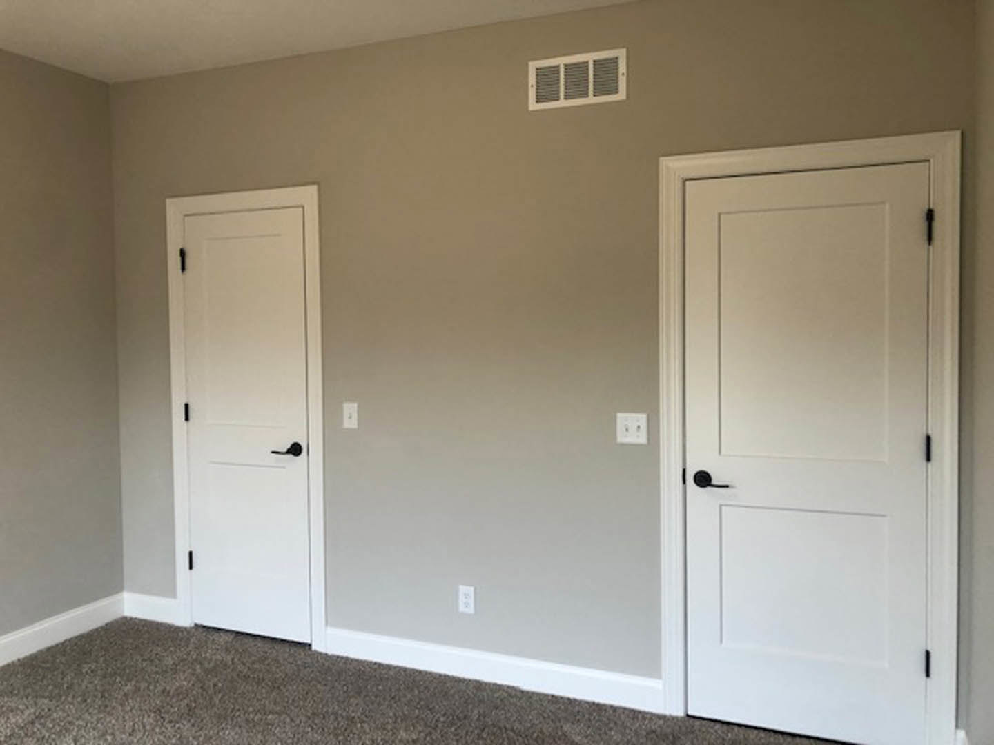 White paneled doors with black handles, square frames, and a wall-mounted vent and light switch on smooth plaster walls above light wood flooring
