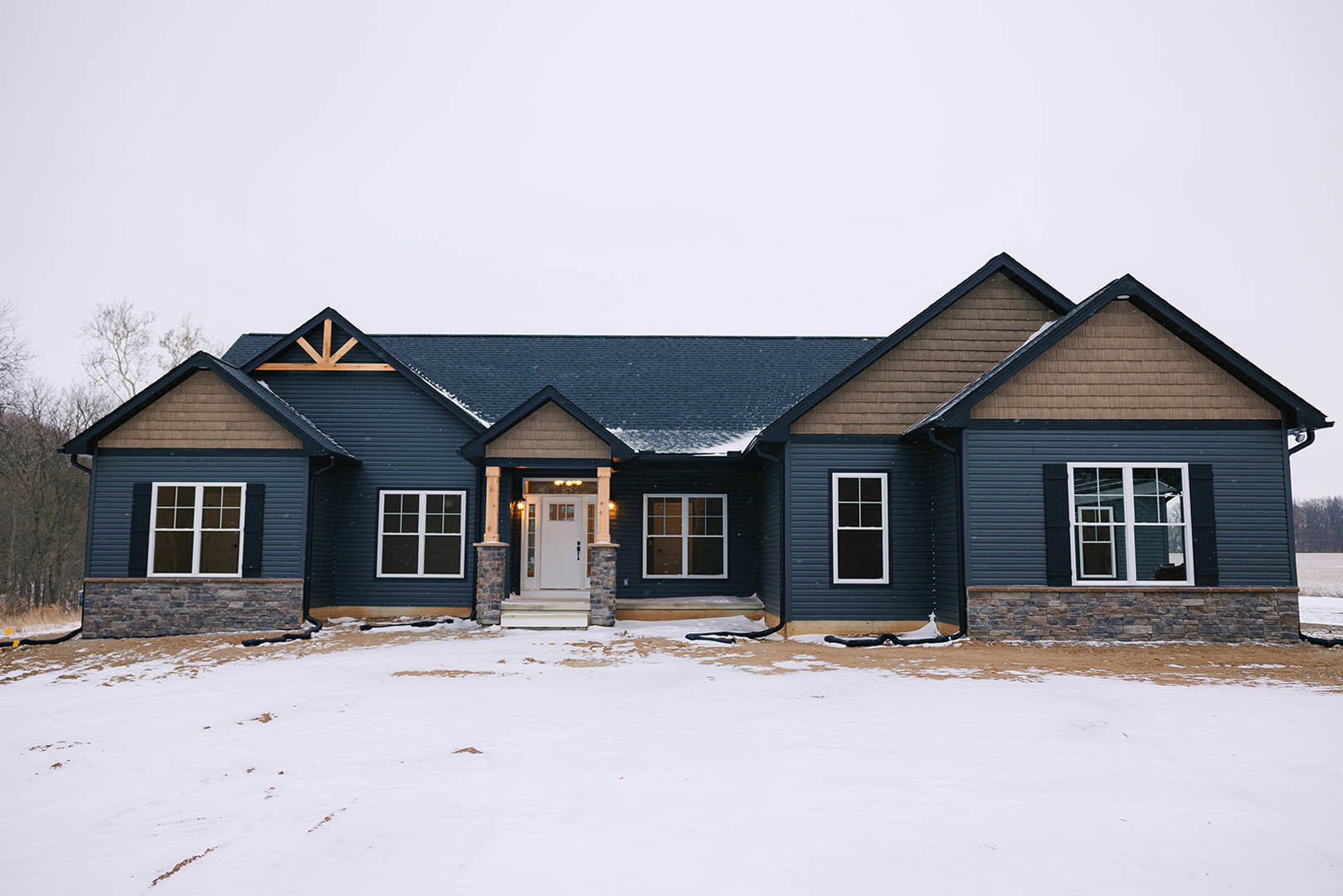 Two-story house with gray siding, white trim, and white front door, snow covering the ground, black pipe visible near foundation, large windows with white frames