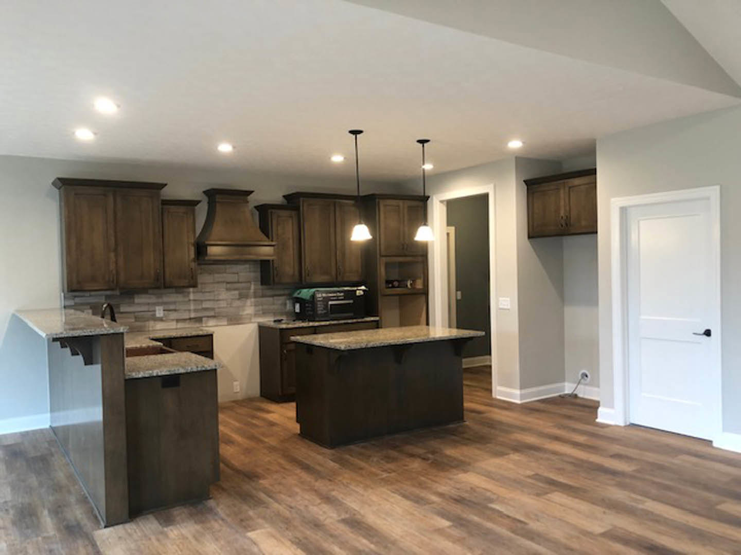 Kitchen with natural wood cabinets, matching wood flooring, white countertop island with built-in sink, white door featuring black handle, and modern appliances.