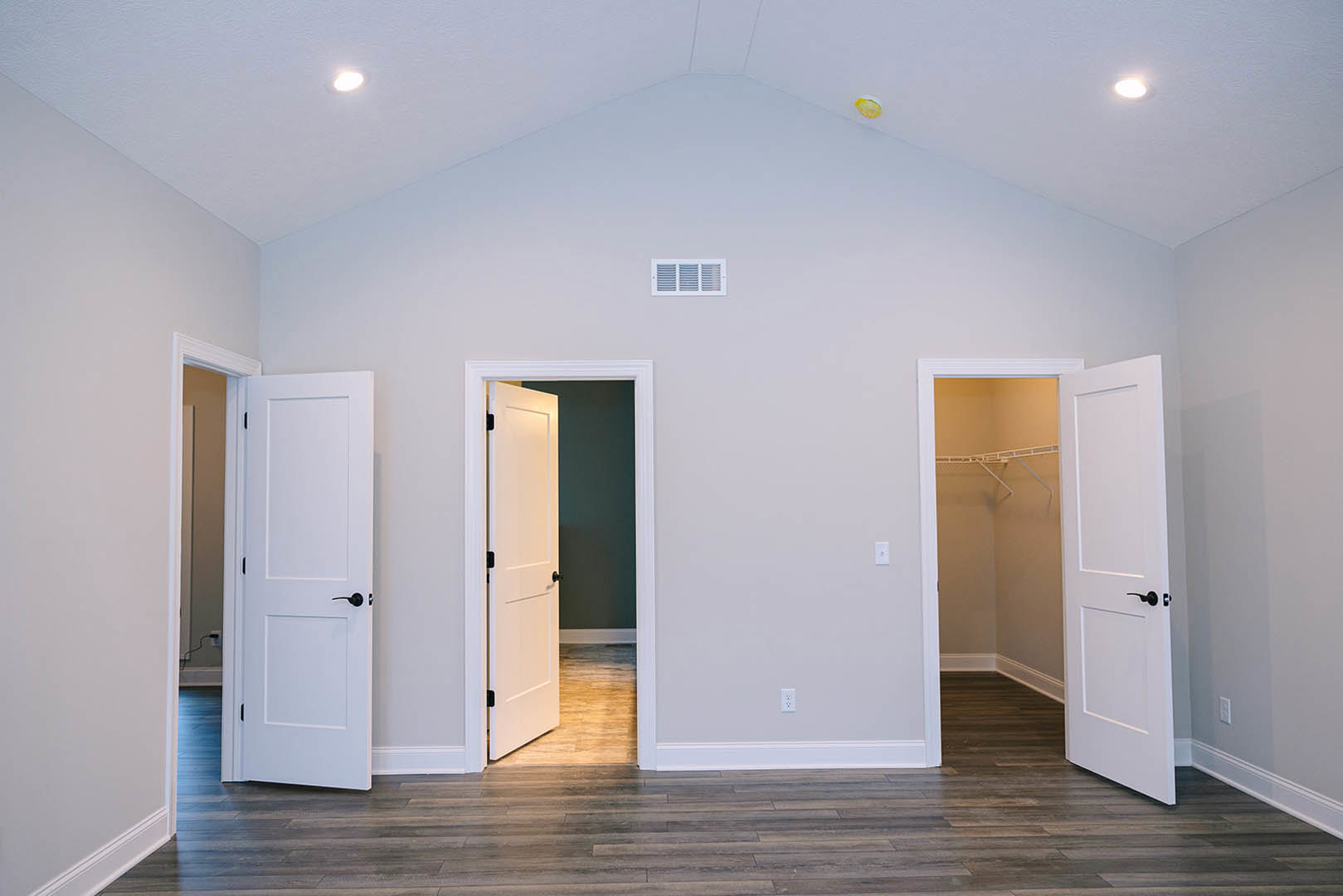 White paneled door with black handle, wood floor, white baseboard, and wall vent in a bright interior room