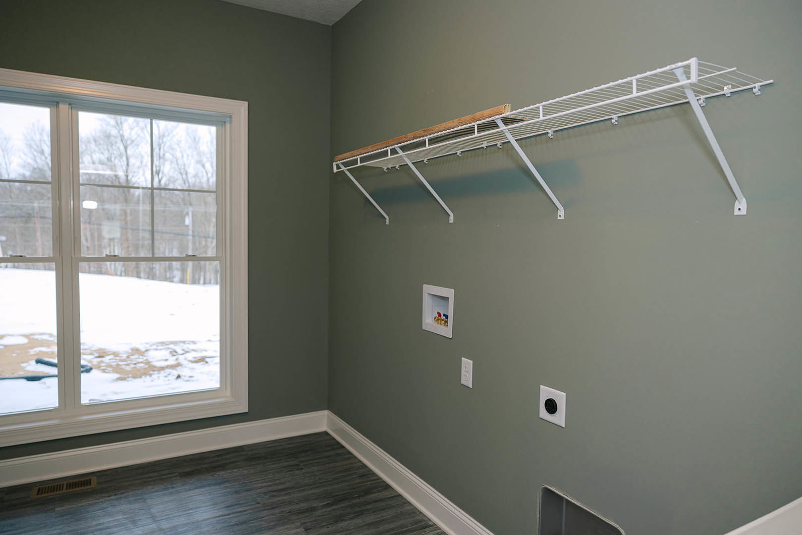 White-walled room featuring a dark wood floor with white baseboard, a white shelf holding exposed valves and a wood piece, large window overlooking snowy landscape, and close-up of
