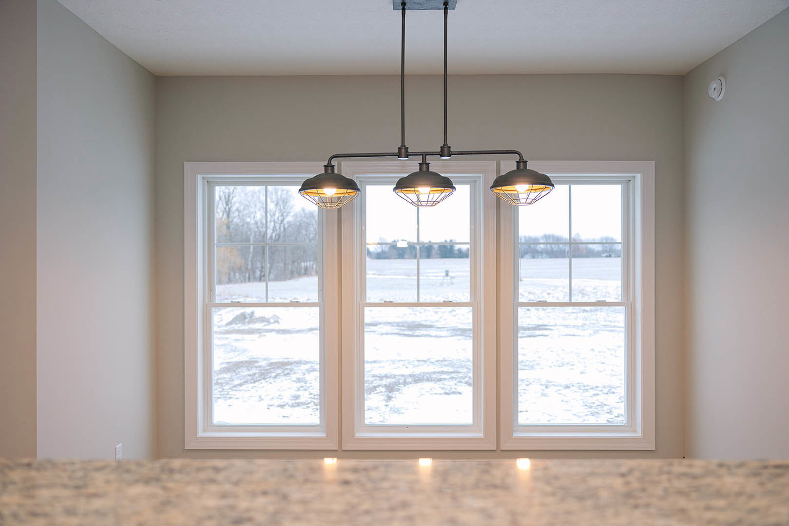 Wire cage ceiling light fixture hanging in a room with large windows, white walls, and window blinds.