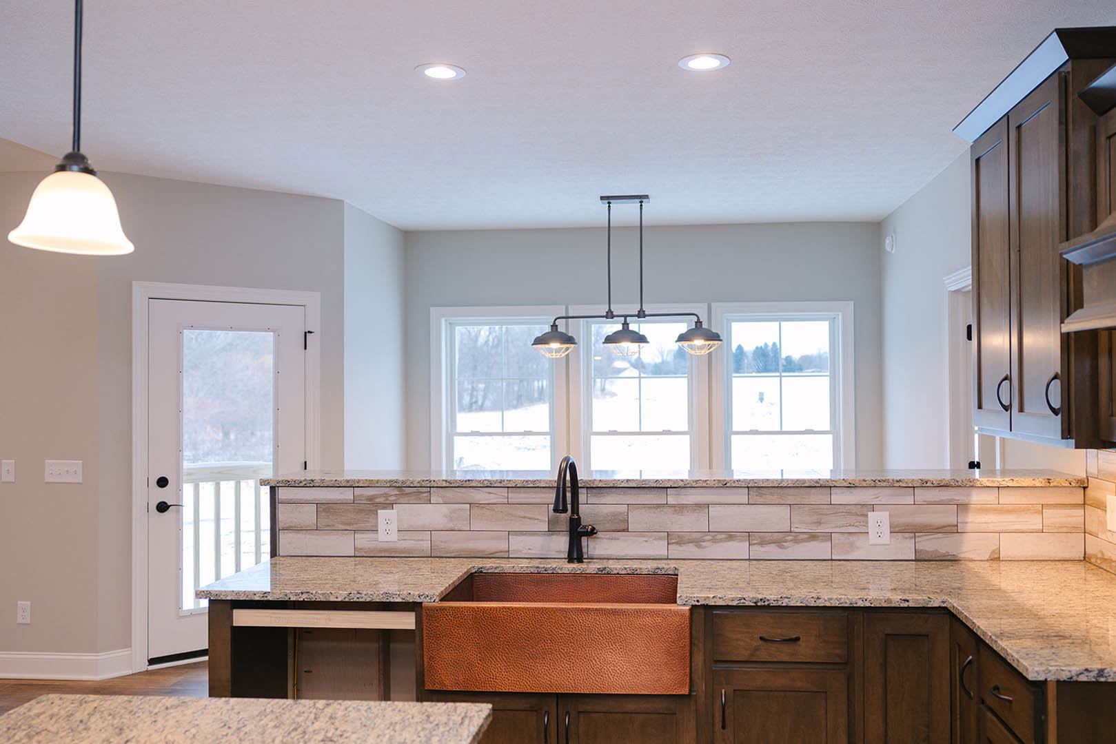 Marble countertop with undermount sink, brown wood cabinetry, glass door, tile backsplash, stainless steel faucet, pendant light fixture