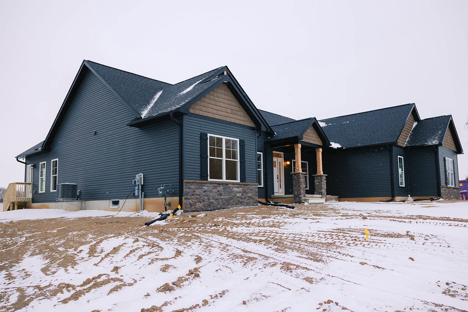 Two-story house with stone and siding exterior, covered porch, wooden deck with railing, tire tracks in snow-covered yard, large windows, winter sky overhead