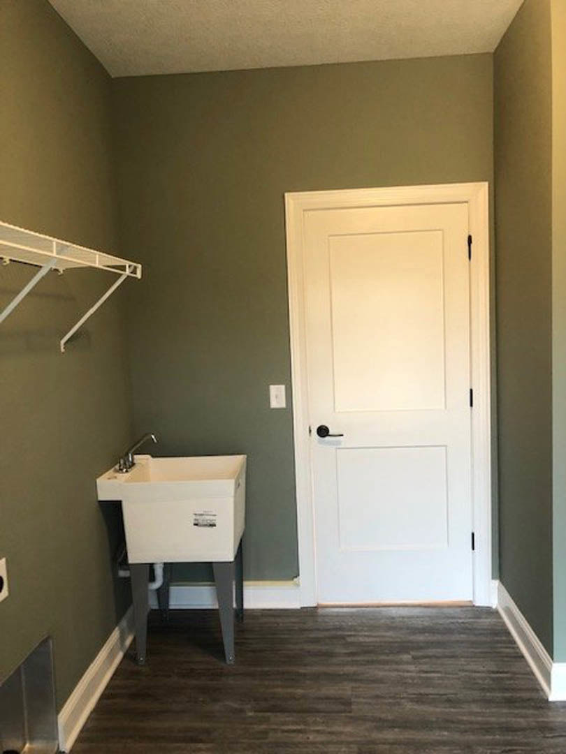 White door with black handle beside a white sink, wood flooring, white shelf with metal rods, bathroom interior.
