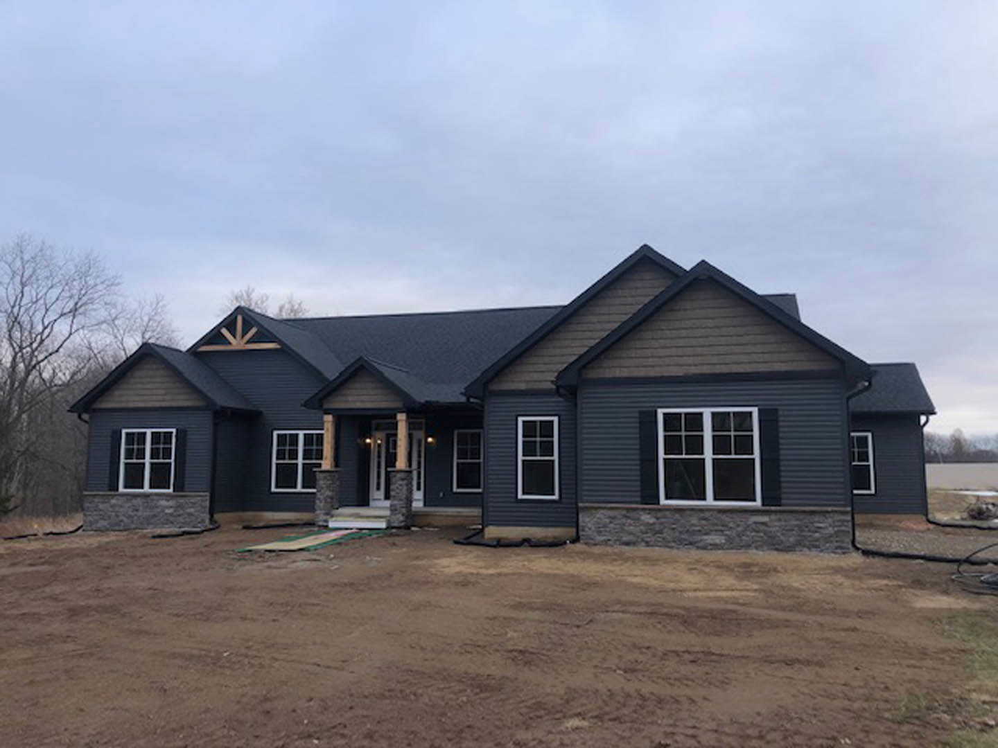 Two-story house with white-framed windows, covered porch, and brown dirt yard; mature tree and cloudy sky in background.