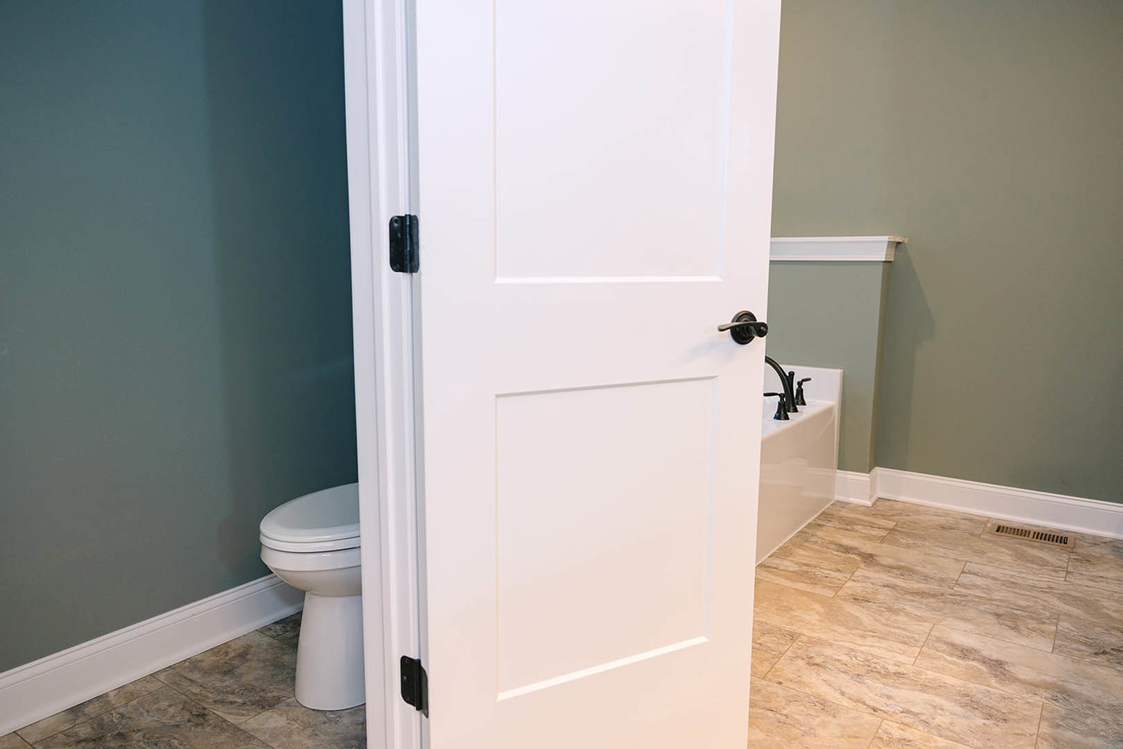 White door with black handle beside white bathtub and toilet, tile floor, light-colored bathroom walls