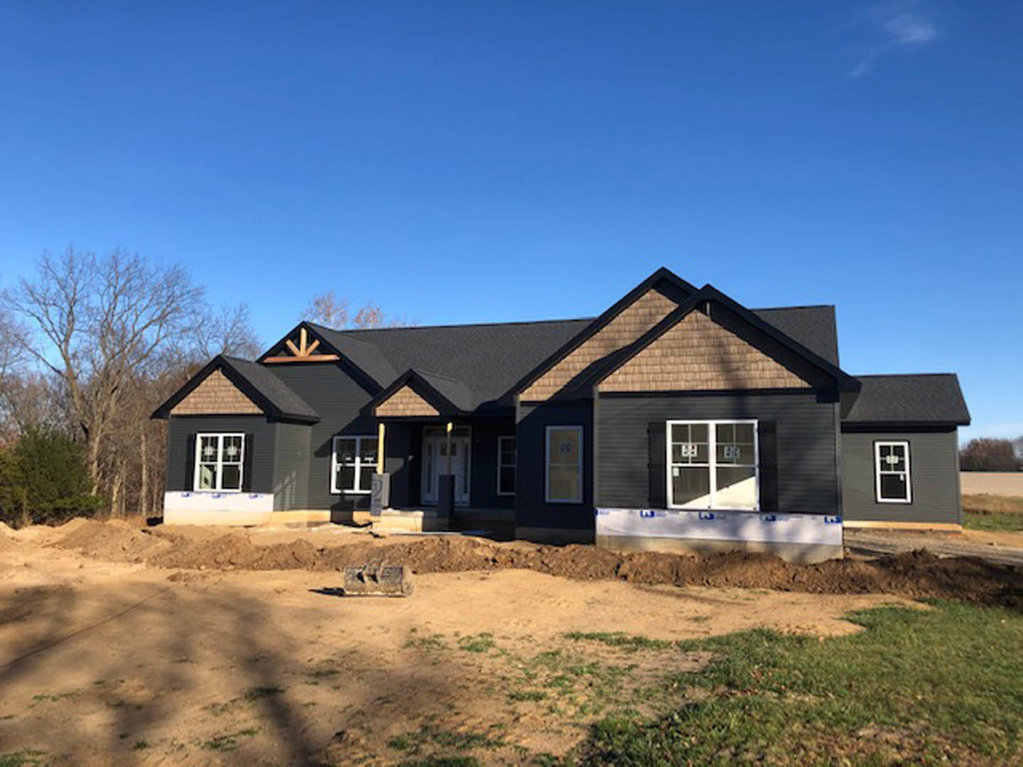 Partially built house with exposed framing, surrounded by dirt and mature trees, under clear blue sky