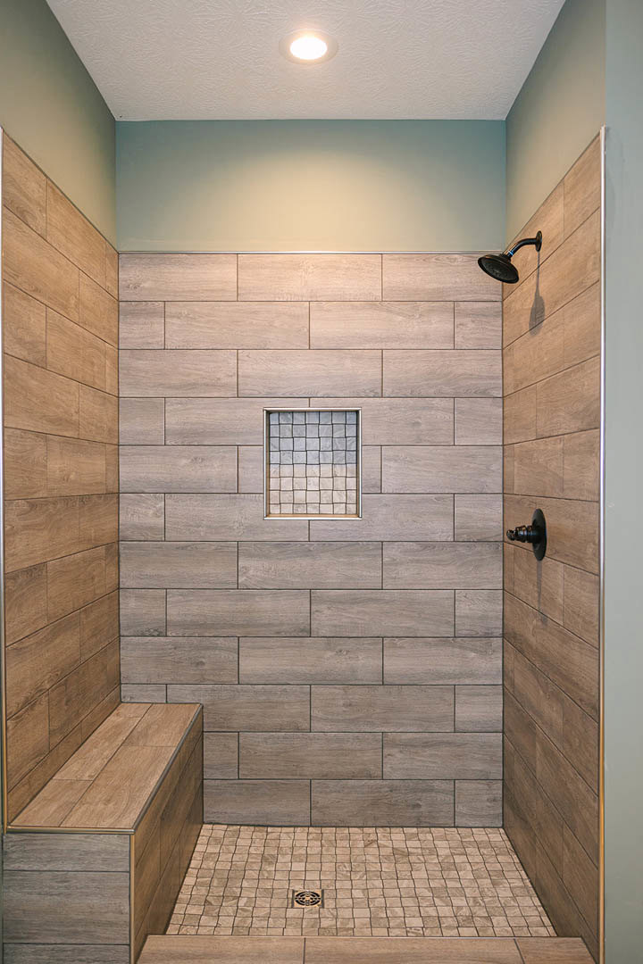 Shower with wood-look tile walls, black shower head, white tile floor with metal drain, square window in tile wall, close-up of metal door knob