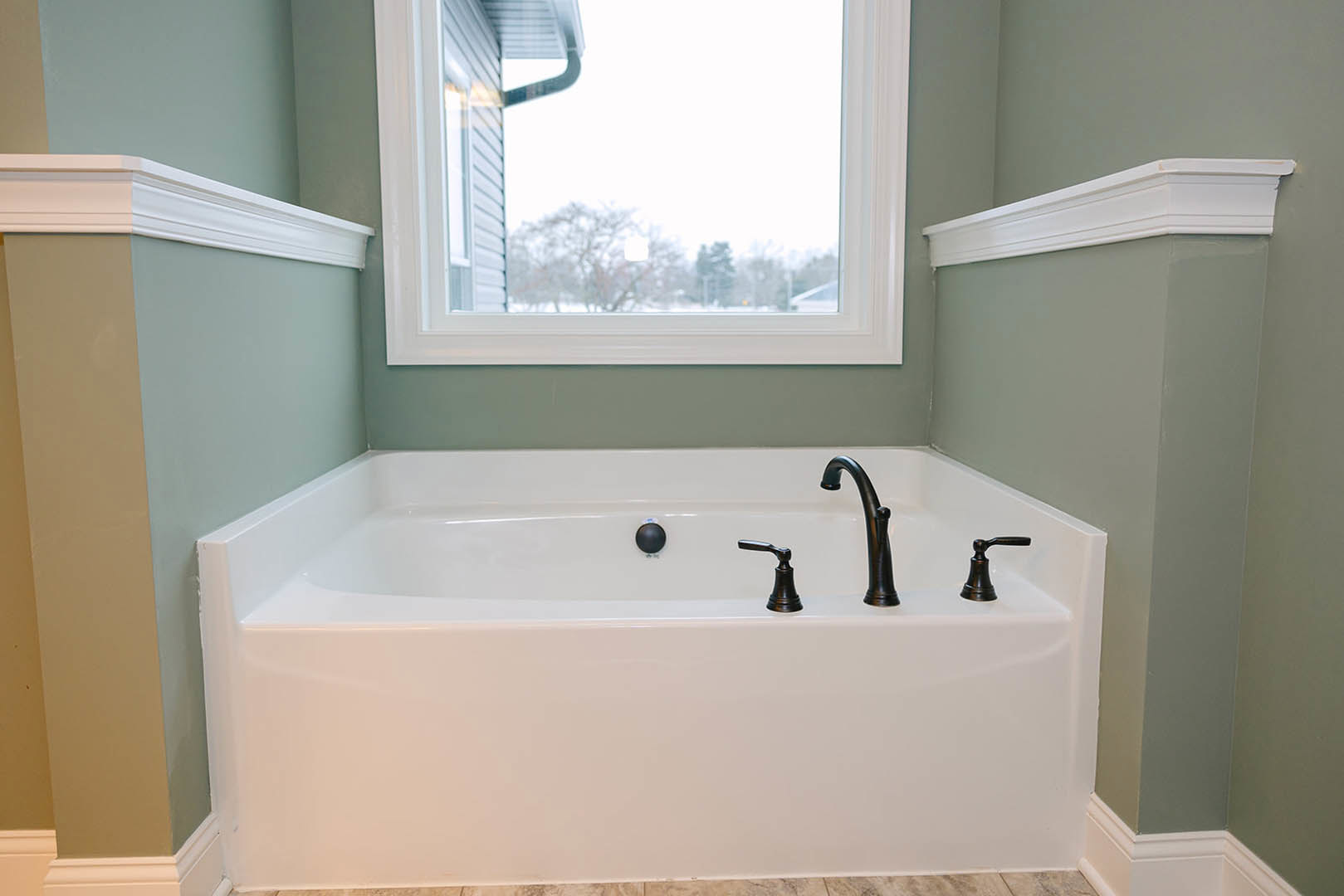 Freestanding white bathtub with chrome faucet beneath large window, light gray walls, natural light illuminating bathroom interior