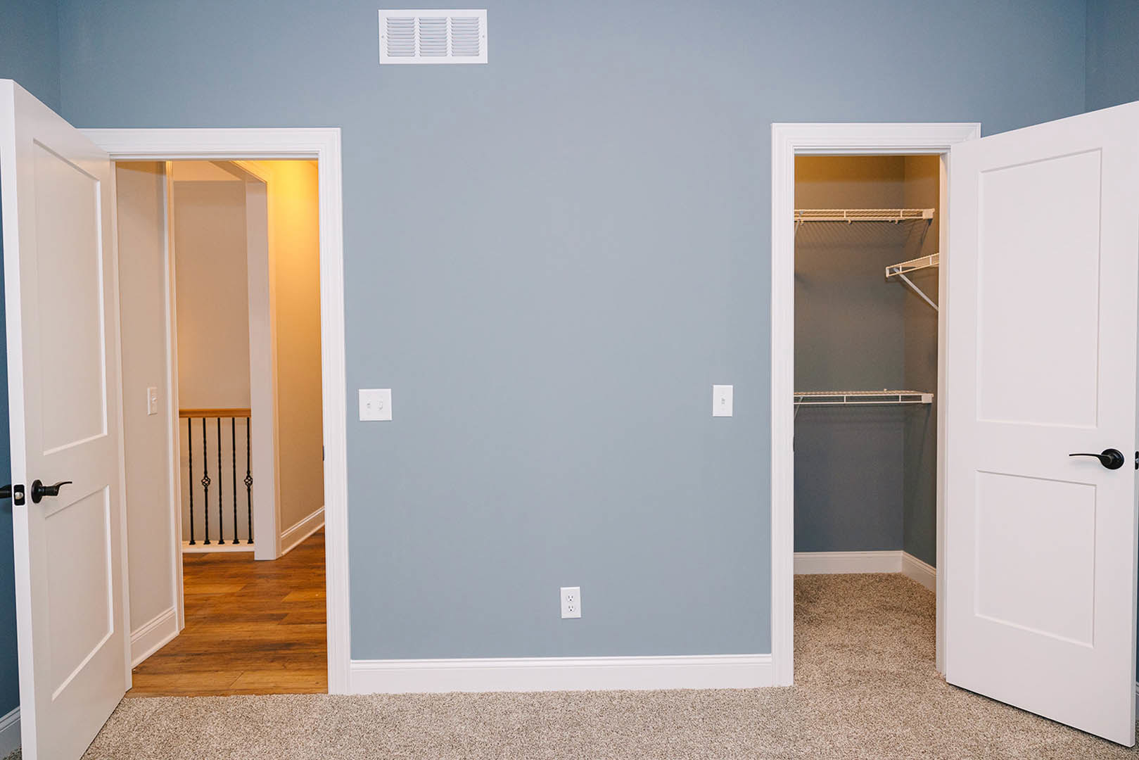 White closet door with black handle, wood floor and white baseboard trim, white vent and light switch on pale wall