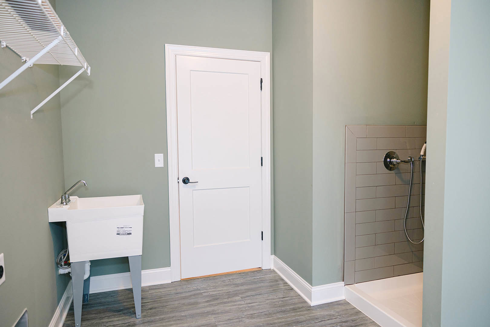 White bathroom with freestanding sink and exposed plumbing, wall-mounted shower head with hose, white bathtub, white door with black handle, and open shelf with metal rods against