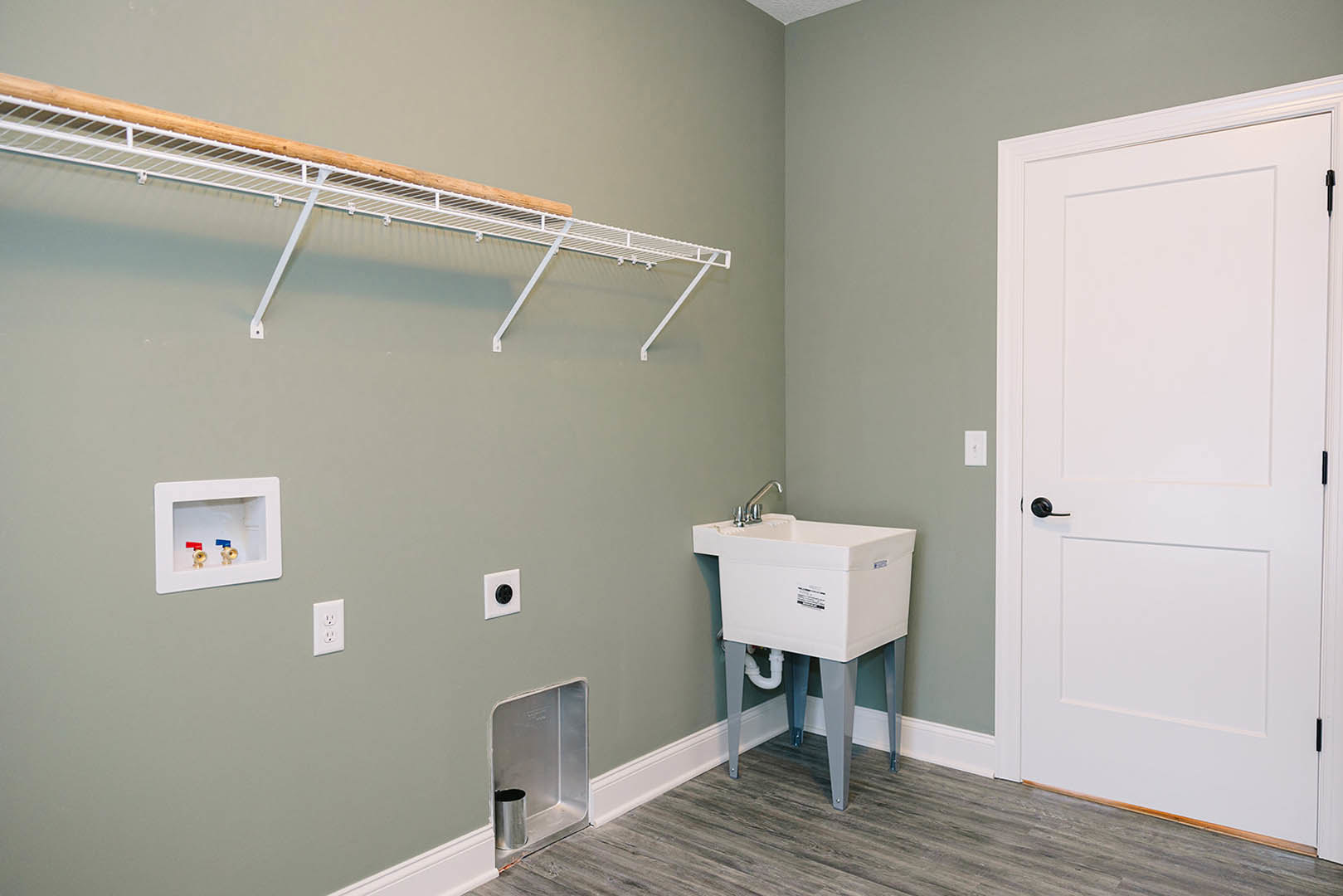 Laundry room with white walls, white door with black handle, freestanding white sink, wall-mounted plumbing box with two valves, white outlet with black circle, and white shelf