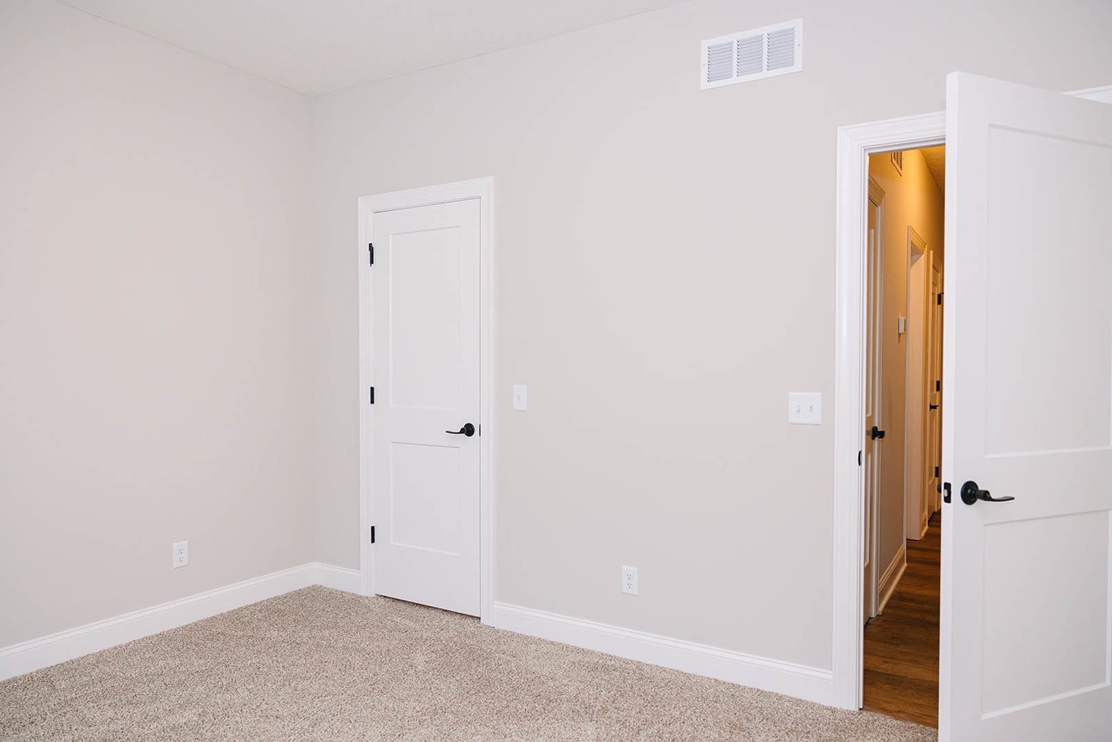 Open white door with black handle leading into room with light wood flooring, white walls, white vent, and white light switch