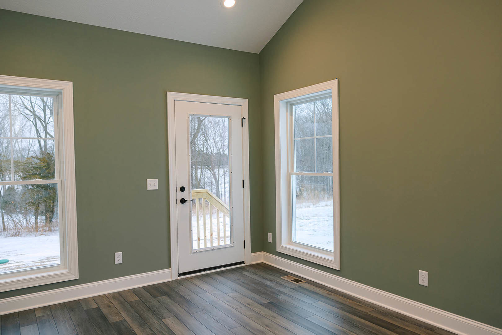 Bright room featuring wood laminate flooring, white walls, a wooden door with glass panels, two large windows overlooking green trees, and a close-up of a wooden railing.