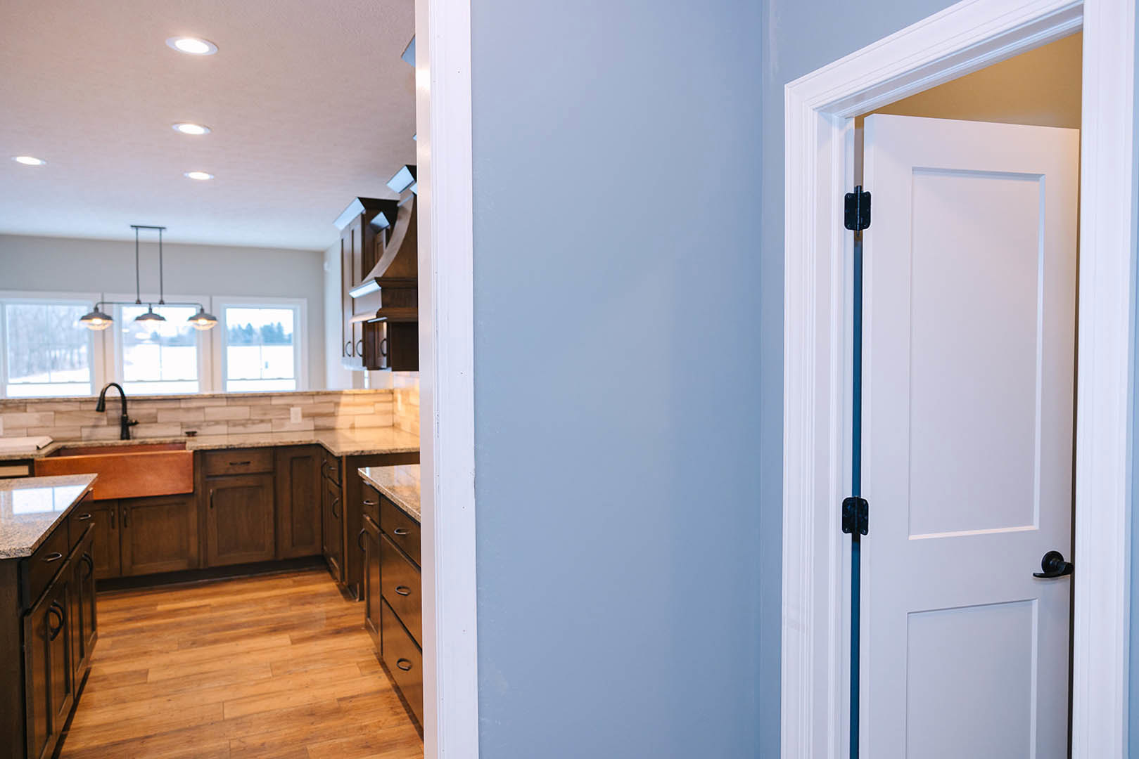 White kitchen door with black handle, wood flooring, white cabinetry, countertop, and sink; light fixture visible in background.