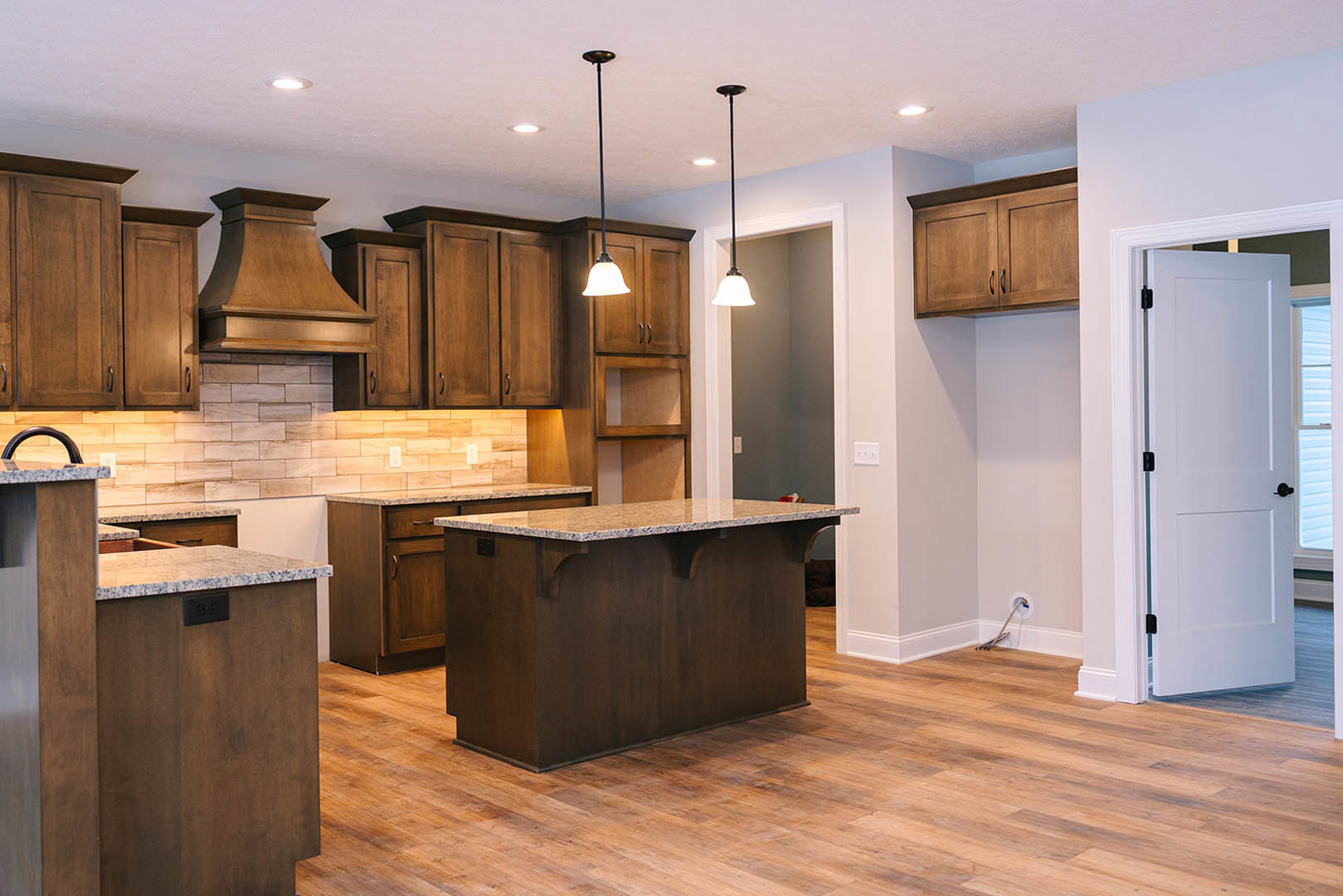 Kitchen with natural wood cabinets and matching wood floor, marble countertop, stainless steel sink, white door, wood range hood, recessed lighting, and visible wiring near wall.