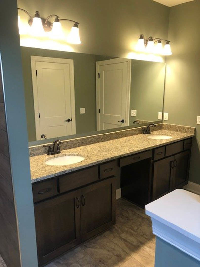 Bathroom with double sinks set in a white countertop, black faucets mounted on a stone tile wall, white cabinetry with black handles, three-bulb light fixture above, and a toilet