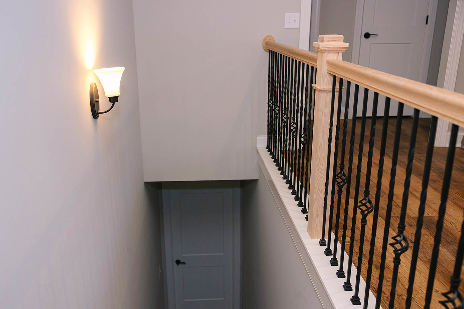 White plaster staircase with black metal handrail, wall-mounted sconce light, white door with black handle, and light switch on adjacent wall