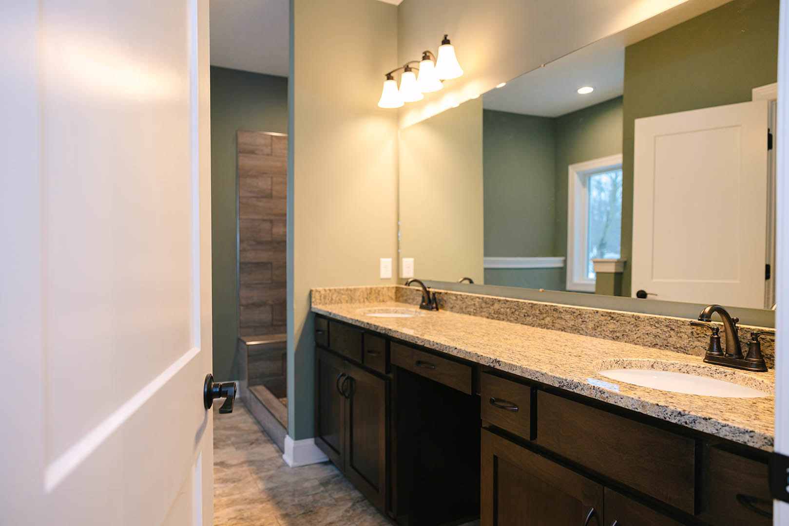 Bathroom with granite countertop, wide mirror above sink, chrome faucet, white door, and multi-bulb light fixture; window visible in background.