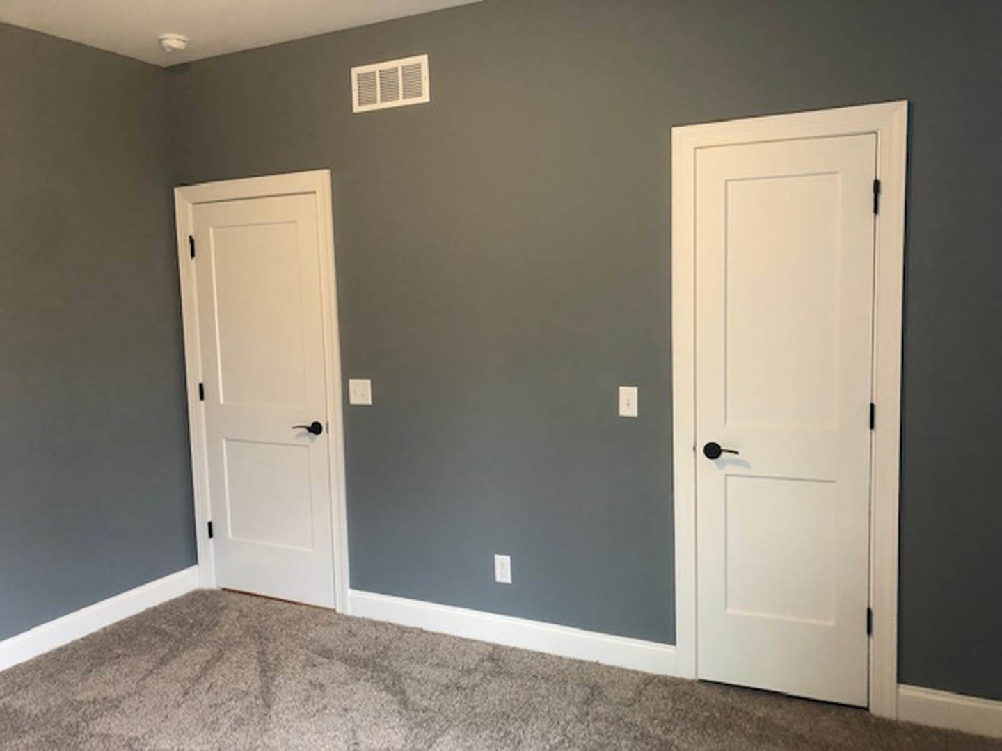 White paneled doors with black handles set in a room with white walls, black trim, and light carpet flooring; wall vent visible near the baseboard.