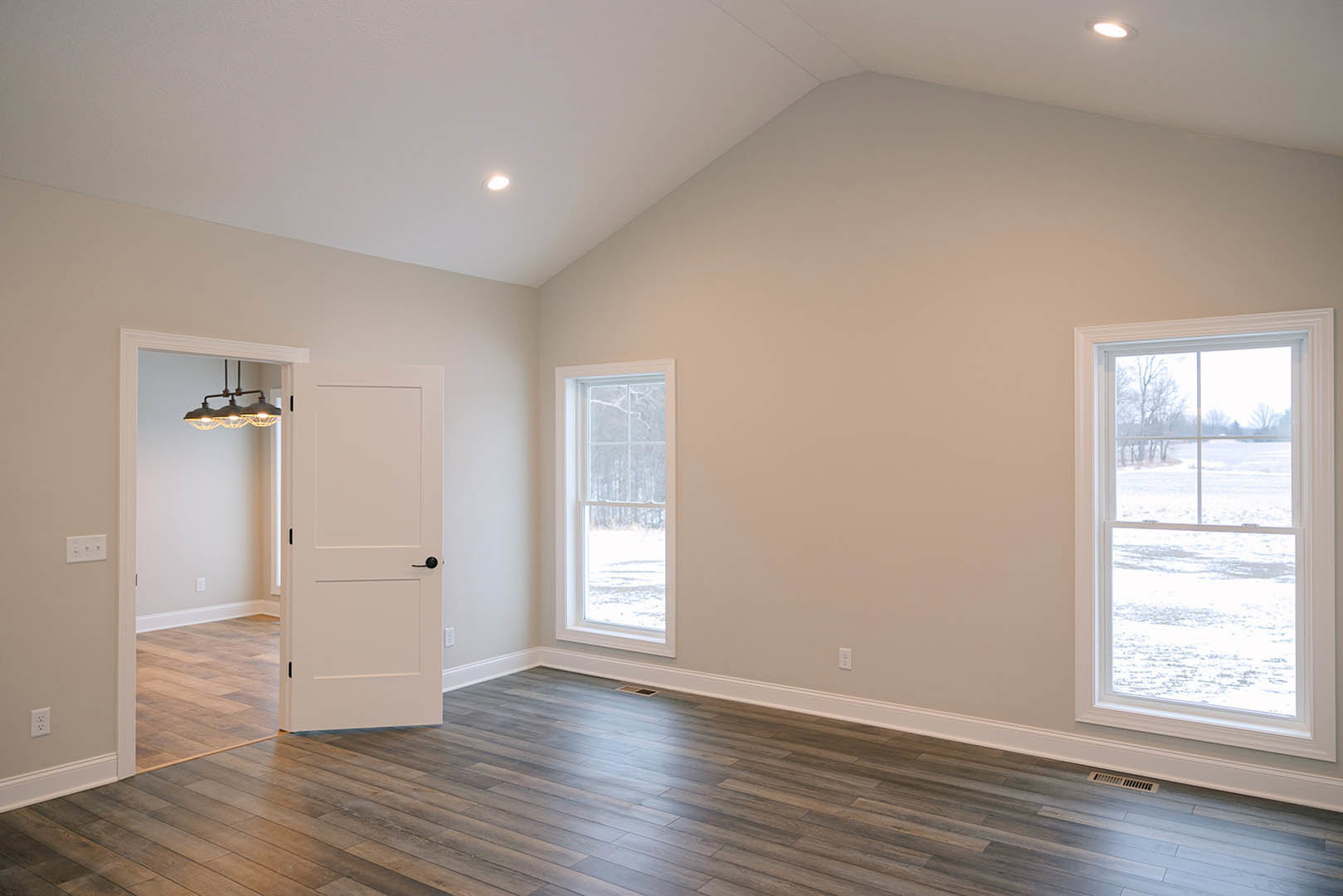 White paneled door with black handle, large windows overlooking snowy field, wood flooring with wall vent, close-up of modern light fixture, smooth plaster walls