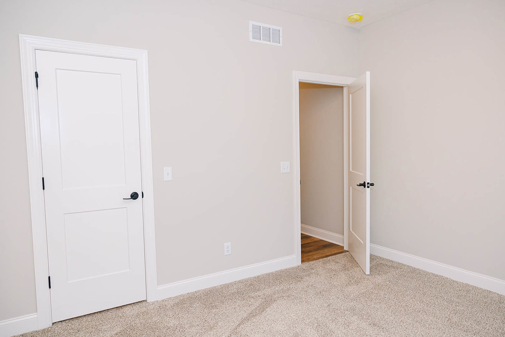 Open white door with black handle leading into carpeted room, white wall vent visible, transition to wood flooring in foreground