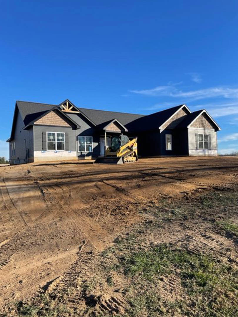 Yellow bulldozer parked on dirt driveway in front of two-story house with white-framed windows, gray siding, and gabled roof under partly cloudy sky