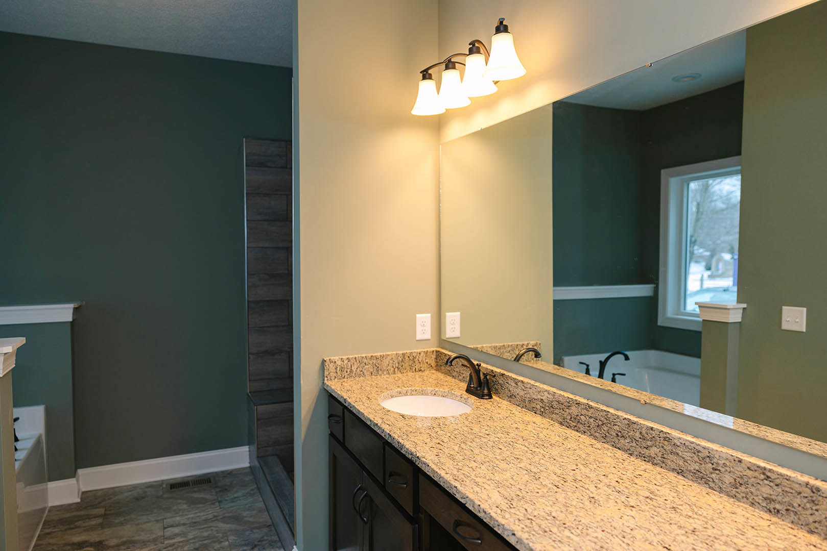 Bathroom with green walls, large mirror above a countertop with sink, white light switch, modern light fixture, window overlooking snowy landscape, bathtub, and black rectangular
