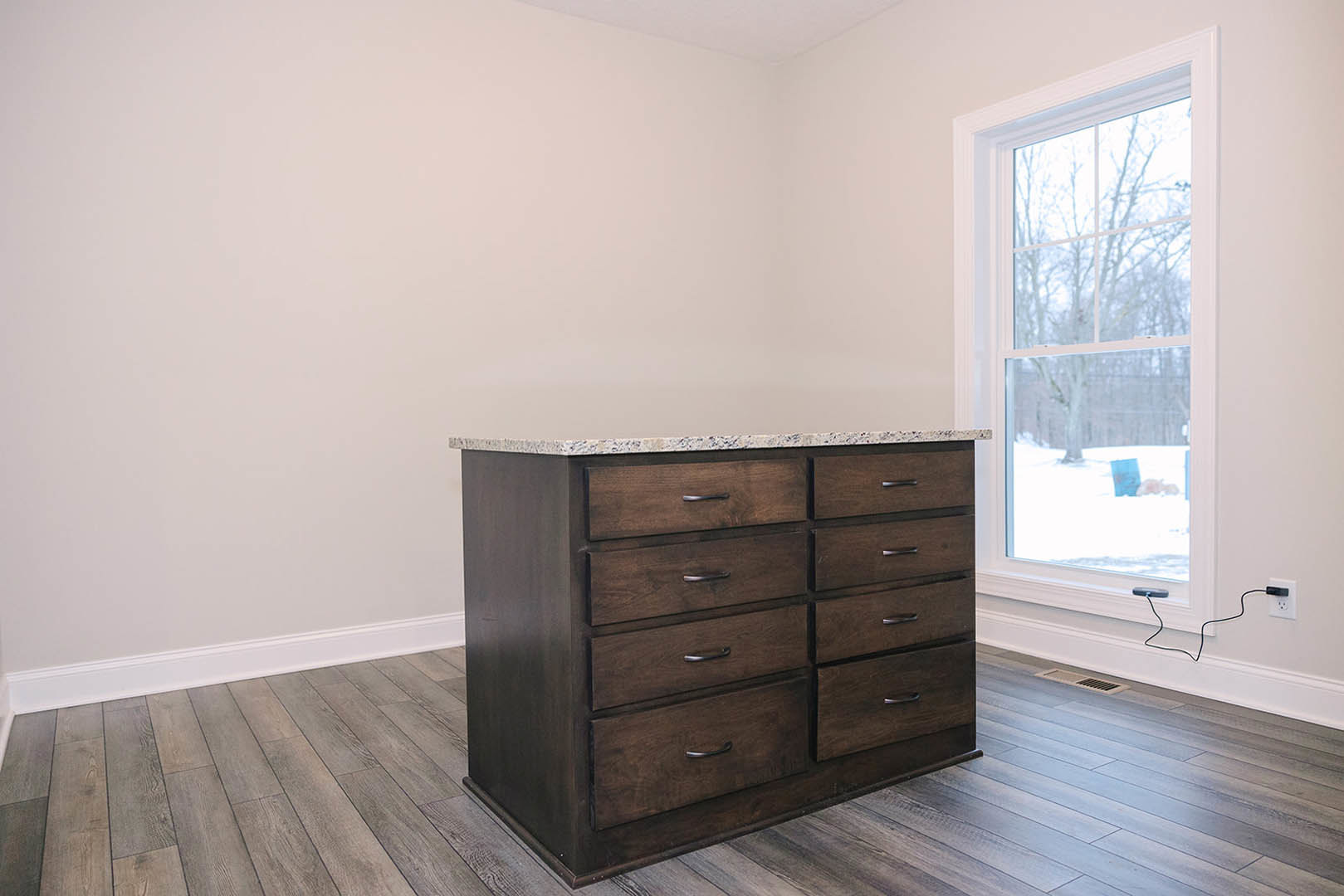 Wooden dresser with multiple drawers against a white wall, black cord plugged into outlet, window overlooking snowy landscape, hardwood floor