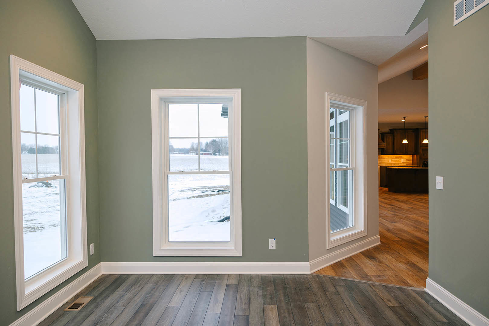 Sunlit room featuring large windows overlooking a snowy field, wood flooring with white baseboards, and a floor vent near the window.
