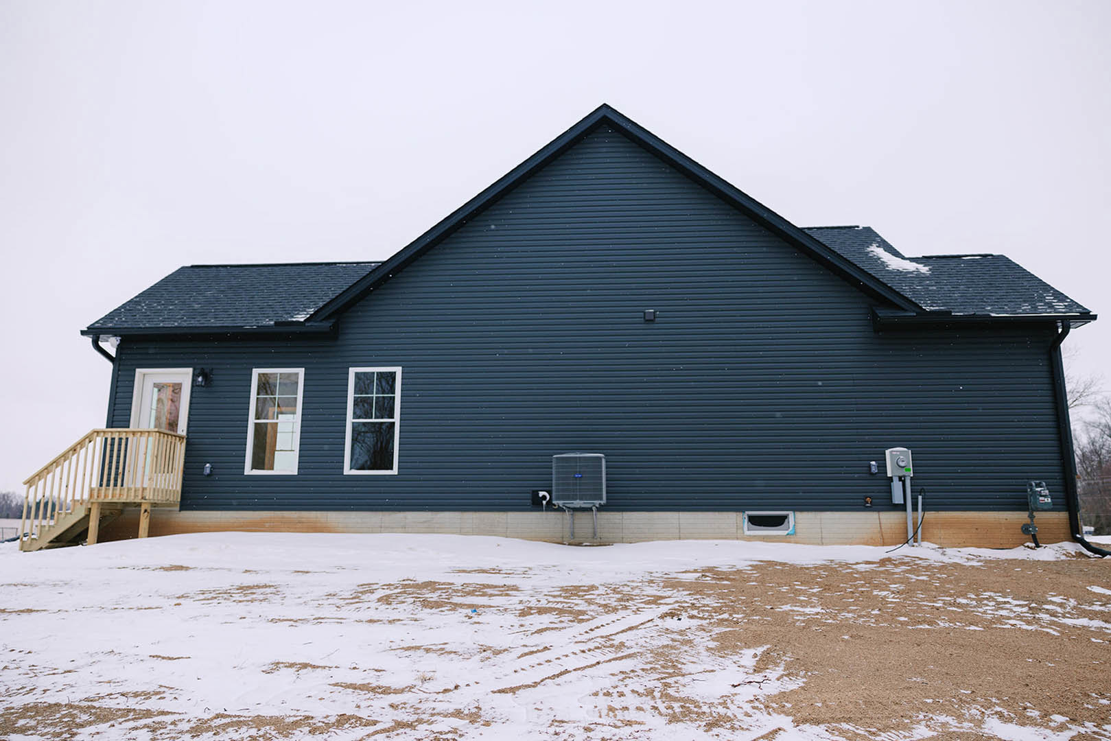 Wood siding house with white-framed window, wooden deck and railing, snow-covered ground with visible tire tracks, winter sky in background