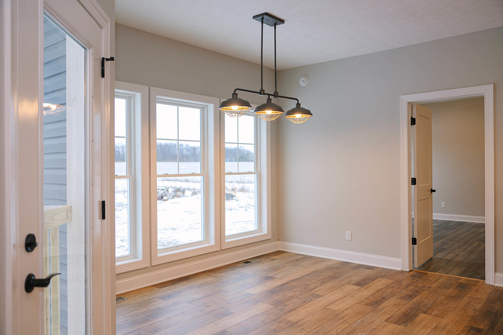 Wood flooring with white baseboards, ceiling-mounted light fixture, white door, large windows overlooking a snowy field