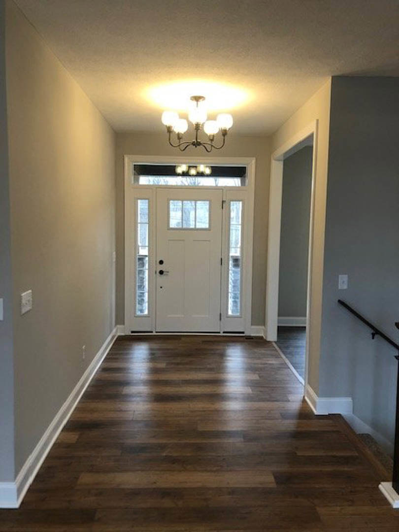 Hallway with hardwood flooring, white door featuring glass window panes, ornate chandelier overhead, and tree visible through window.